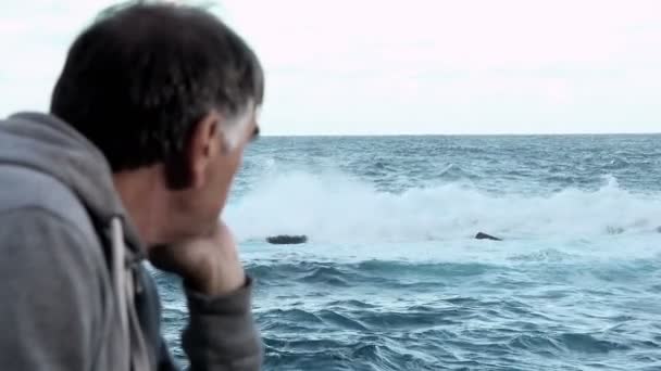 Hombre maduro observa las olas estrellándose sobre las rocas — Vídeo de stock