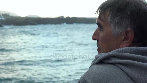 Зрелый мужчина расслабляется, наблюдая за морем в облачный день — стоковое видео