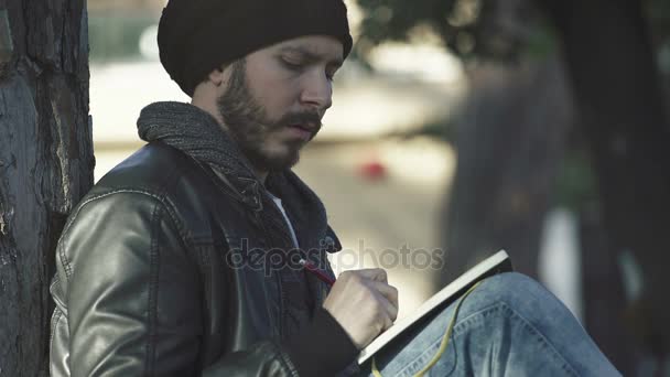 Молодой человек, сидящий под деревом с намерением написать в блокноте — стоковое видео