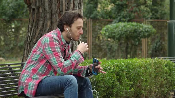 Молодий чоловік сидить на лавці, сперечаючись тепло по телефону — стокове відео