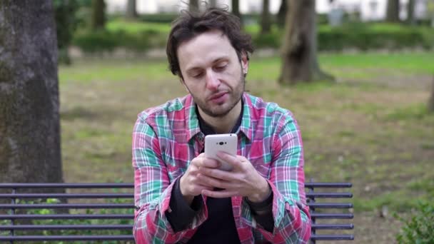 Влюбленный мужчина, сидящий в парке, пишет сообщение на своем смартфоне своей девушке — стоковое видео