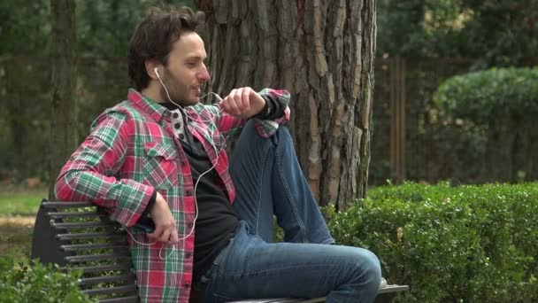 Расслабленный мужчина в парке, разговаривает по телефону с наушниками — стоковое видео