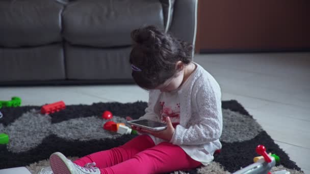 Niña sentada en el suelo con la intención de jugar con el teléfono inteligente — Vídeos de Stock