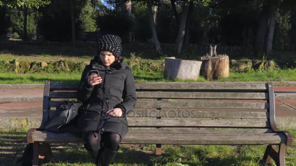 Jovem senhora sentado em um banco lê uma mensagem no smartphone — Vídeo de Stock
