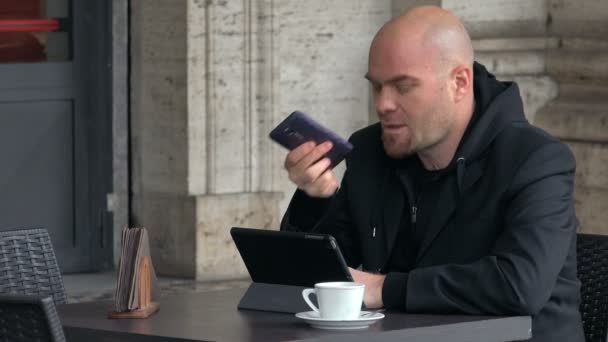 Ocupado careca falando ao telefone e assistindo em tablet sentar na mesa — Vídeo de Stock