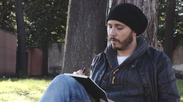 Bärtiger Mann sitzt unter einem Baum und schreibt in ein Notizbuch — Stockvideo