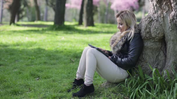 Blondine sitzt mit Tablet unterm Baum — Stockvideo