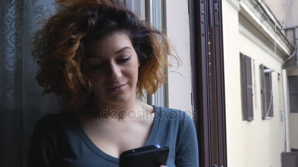 リラックスしたセクシーな女性は、開いた窓の近くのスマート フォンでメッセージを書き込みます — ストック動画