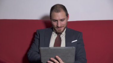 Tablet kullanarak kırmızı kanepede oturan işadamı gülümseyen