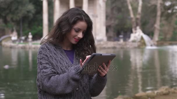 Uśmiechający się piękna kobieta za pomocą tabletu, jezioro park w tle — Wideo stockowe