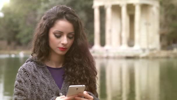 Cantik jenis brunette di smartphone di taman, danau di latar belakang — Stok Video