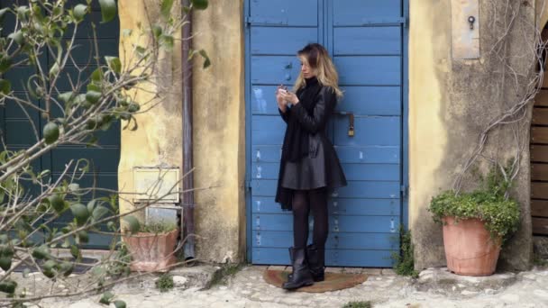 Очаровательная женщина использует смартфон, прислонившись к синим воротам — стоковое видео