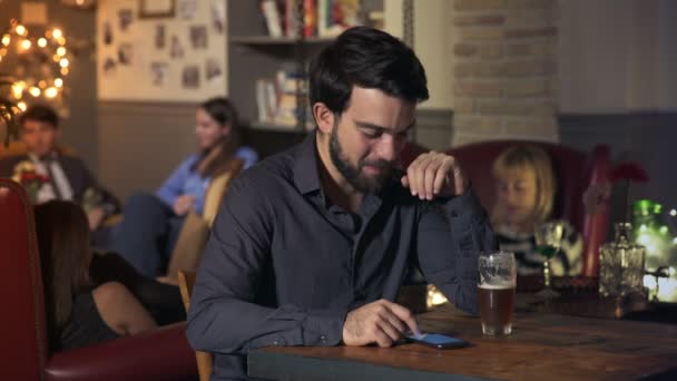 Hombre sonriente sentado en el bar con cerveza de cristal, recibe un mensaje en el teléfono — Vídeos de Stock