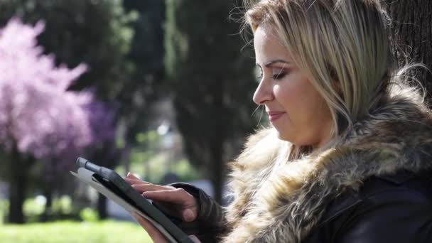 Профиль улыбающейся блондинки в парке, использующей планшет — стоковое видео