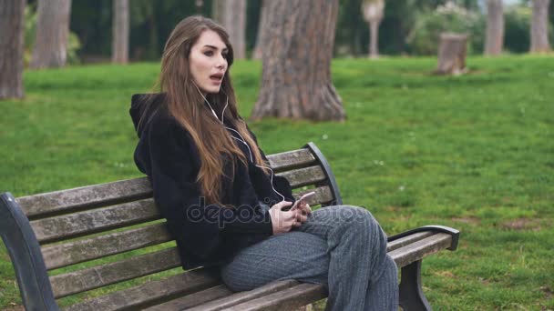 Retrato de una hermosa mujer sentada en el banco mientras escucha música con cabecera — Vídeo de stock