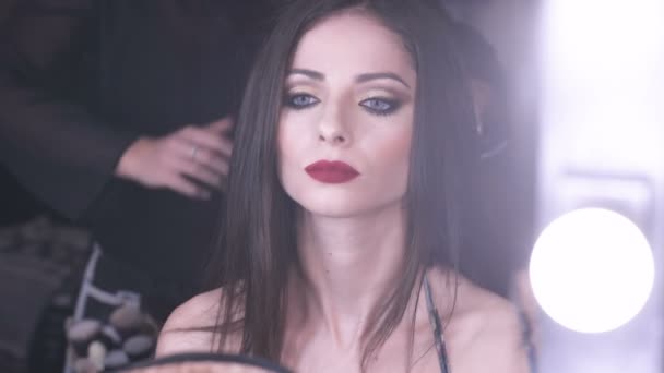 Piękny model wygląda w lustrze, podczas gdy stylista włosów wygładza włosy — Wideo stockowe