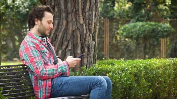 Perfil del hombre sentado en el banco mientras responde a un mensaje con el teléfono inteligente — Vídeos de Stock