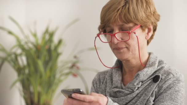 Mujer rubia madura con gafas rojas escribiendo en el teléfono inteligente — Vídeos de Stock