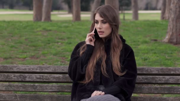 Спокойная женщина, сидящая на скамейке в парке и разговаривающая по телефону — стоковое видео