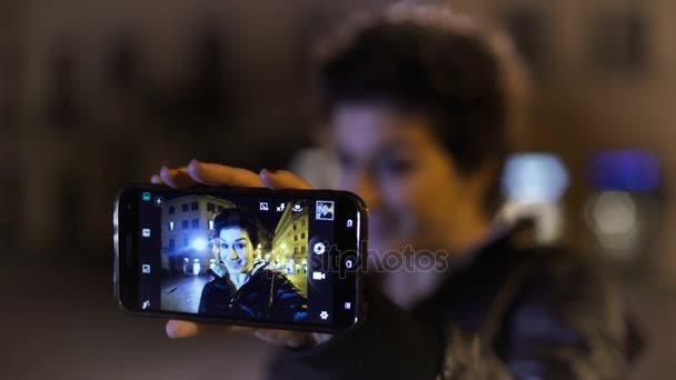 Mujer alegre y sonriente toma selfies en la ciudad por la noche — Vídeo de stock