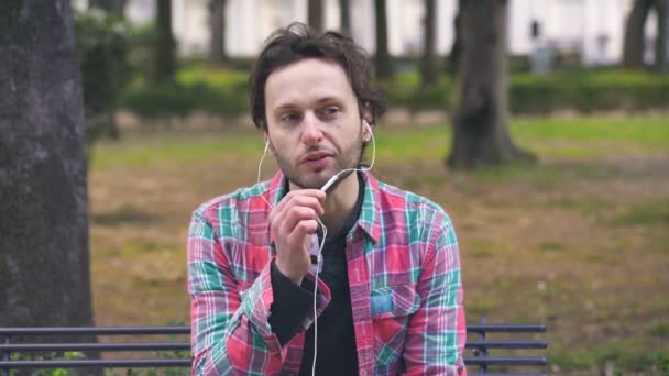 Un uomo affascinante che parla al telefono con le cuffie, seduto su un parco al parco — Video Stock