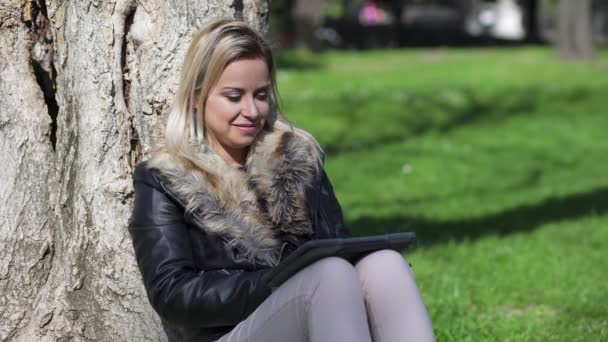 Улыбающаяся молодая женщина сидит под деревом с помощью планшета — стоковое видео