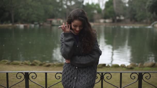 Мила усміхнена молода жінка розмовляє по телефону в парку, на фоні озера — стокове відео
