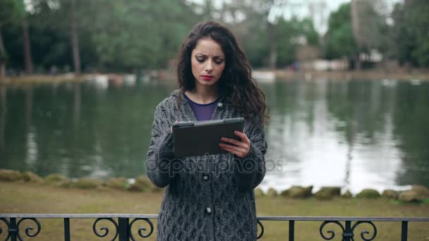 Hermosa morena utiliza la tableta en el parque, lago en el fondo — Vídeos de Stock