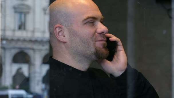 Guapo sonriente calvo hablando por teléfono detrás de una ventana — Vídeo de stock