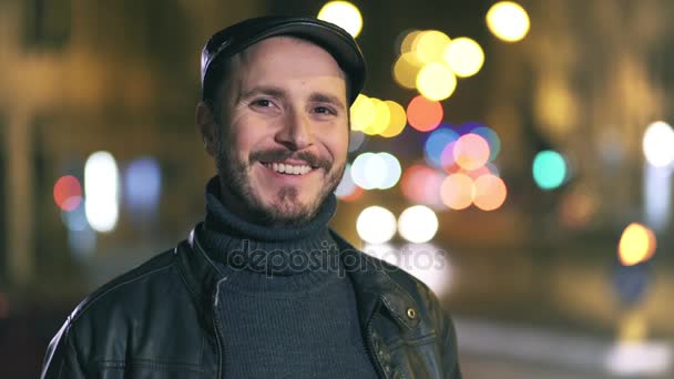 Sorridente uomo affascinante sorridente alla macchina fotografica, luci della città di sfondo di notte — Video Stock