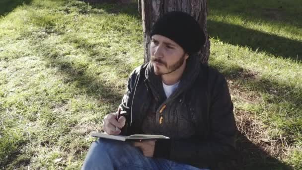 Cher jeune homme assis sous un arbre écrit dans un cahier — Video