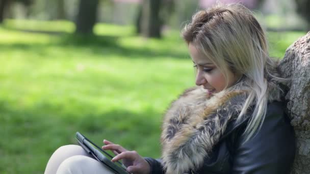 Gülümseyen güzel kadın bir ağacın altında oturup tablet kullanma — Stok video