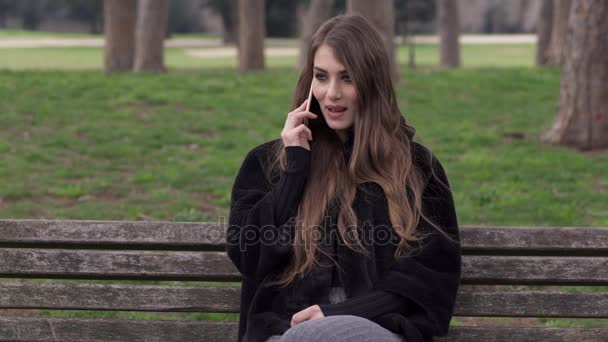 Souriant femme mignonne assis sur un banc de parc parler au téléphone — Video