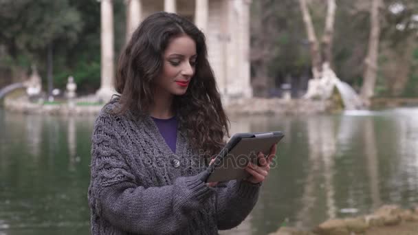 Rilassato sorridente giovane donna utilizzando tablet al parco, lago sullo sfondo — Video Stock