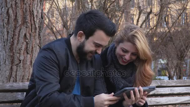Retrato de pareja divertida y sonriente en el banco mirando fotos en la tableta — Vídeos de Stock