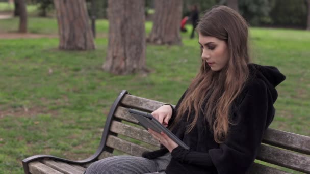 Porträtt av söt ung kvinna skriver på tabletten sitter på bänken i parken — Stockvideo