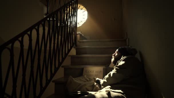 Homeless lying on the twilight stairs looks at the light coming from the window — Stock Video