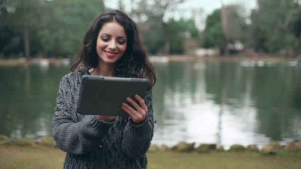 Усміхнена брюнетка використовує планшет в парку, озеро на задньому плані — стокове відео