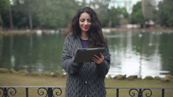 Sonriente, bonita morena utiliza la tableta en el parque, lago en el fondo — Vídeos de Stock