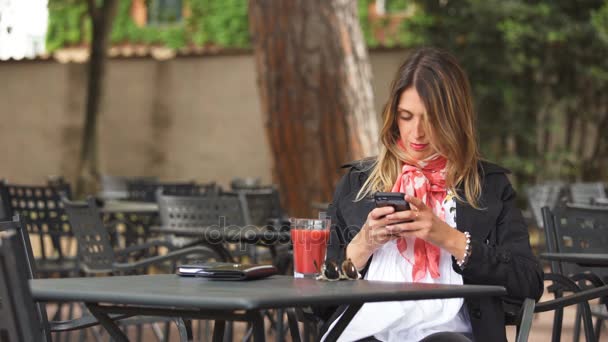 Портрет улыбающейся красивой женщины, которая пишет смс на смартфоне в кафе на открытом воздухе — стоковое видео