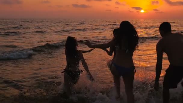 Grupo de cuatro amigos corriendo juntos hacia el mar en una playa al atardecer — Vídeos de Stock
