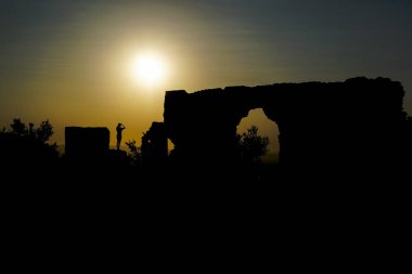 Kadın arka planda harabelerde, ufukta güneş var.