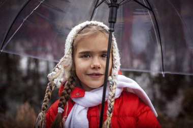 7-8 yaşlarında, kırmızı ceketli, şeffaf şemsiyeli bir kız.