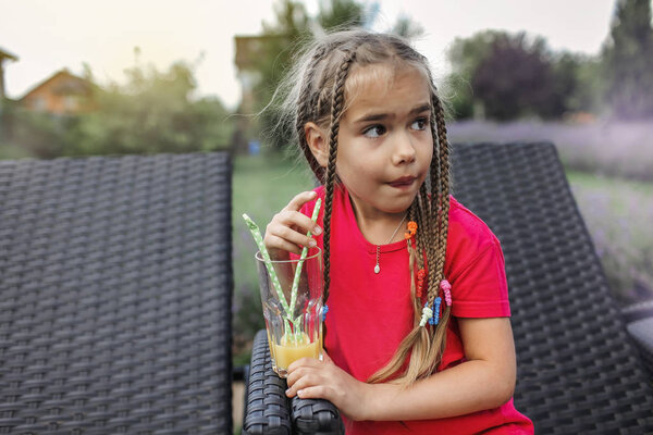 Стильная милая девушка с косичками в синих футболках, пьющая свежий апельсиновый сок с бумажной соломинкой на заднем дворе летнего домика. Витамины и здоровое питание для детей, счастливое детство и летнее время на свежем воздухе
