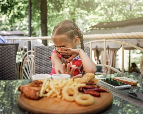 Cute girl sitting at table in open terrace of street cafe and does not want to eat different fried food. Deep-fried cheese, French fries, mushrooms, squid rings with sauces, outdoor summer lifestyle