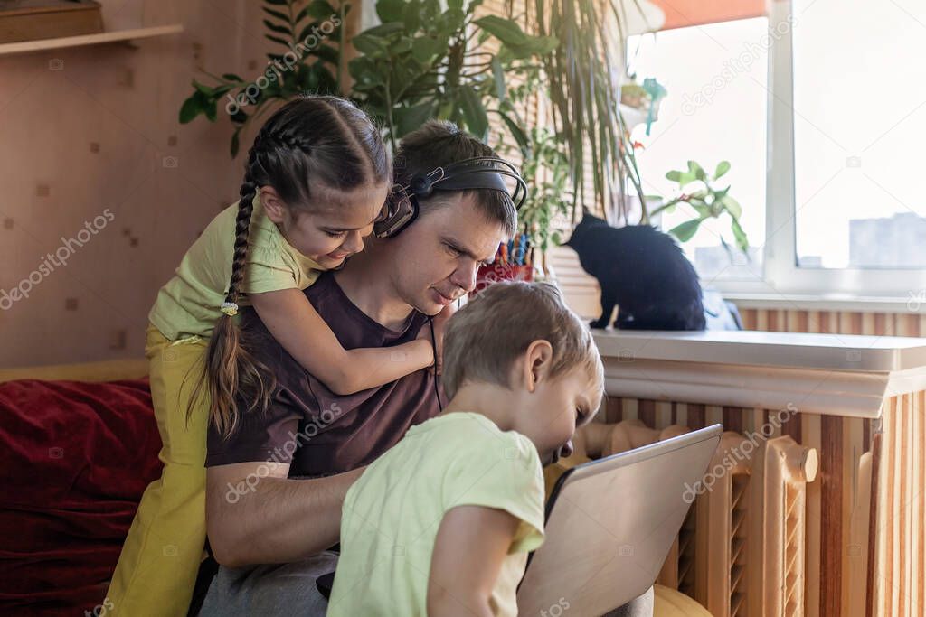 Happy man with joyful children using laptop and earphone during his home working while sitting on sofa at home, home office with together with children, life during quarantine