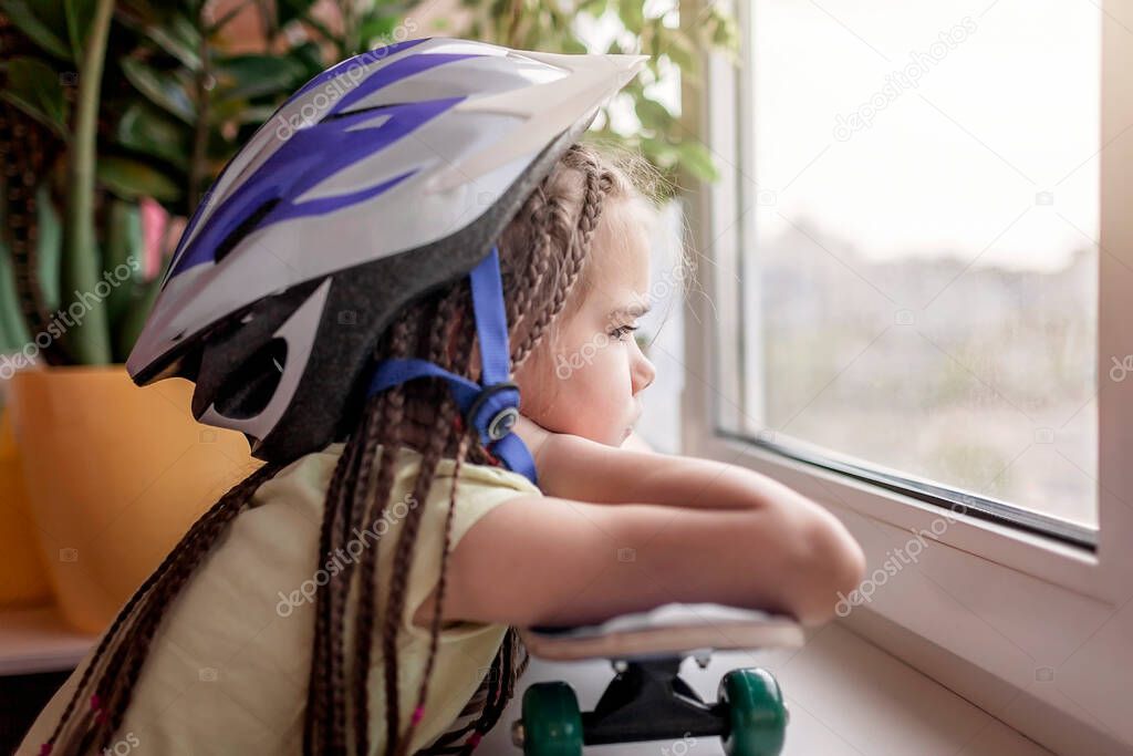 Cute sad little kid sitting on the window sill and looking on the street, dream about outdoor activity during quarantine lockdown, indoor lifestyle, concept of sorrow