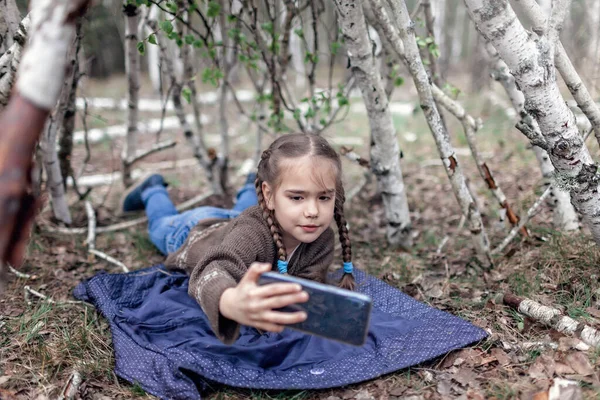 Young girl chatting on mobile phone in the built wooden stick hut house in the wild birch forest during her social distant walking in lockdown time, walking on fresh air, outdoor active lifestyle