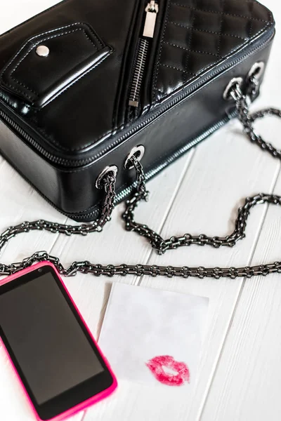 Bolso de mano de cuero negro con cadena sobre fondo de madera blanca, teléfono rosa y un cuaderno . Imagen De Stock