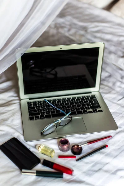 Ordenador portátil, gafas, teléfono y cosméticos se encuentran en una cama blanca . Imágenes De Stock Sin Royalties Gratis